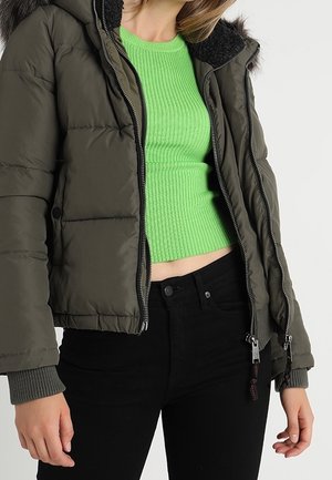 Frau trägt eine aufgeknöpfte dunkelgrüne Steppjacke über einem hellgrünen gerippten kurzen Pullover und schwarze hoch taillierte Jeans.