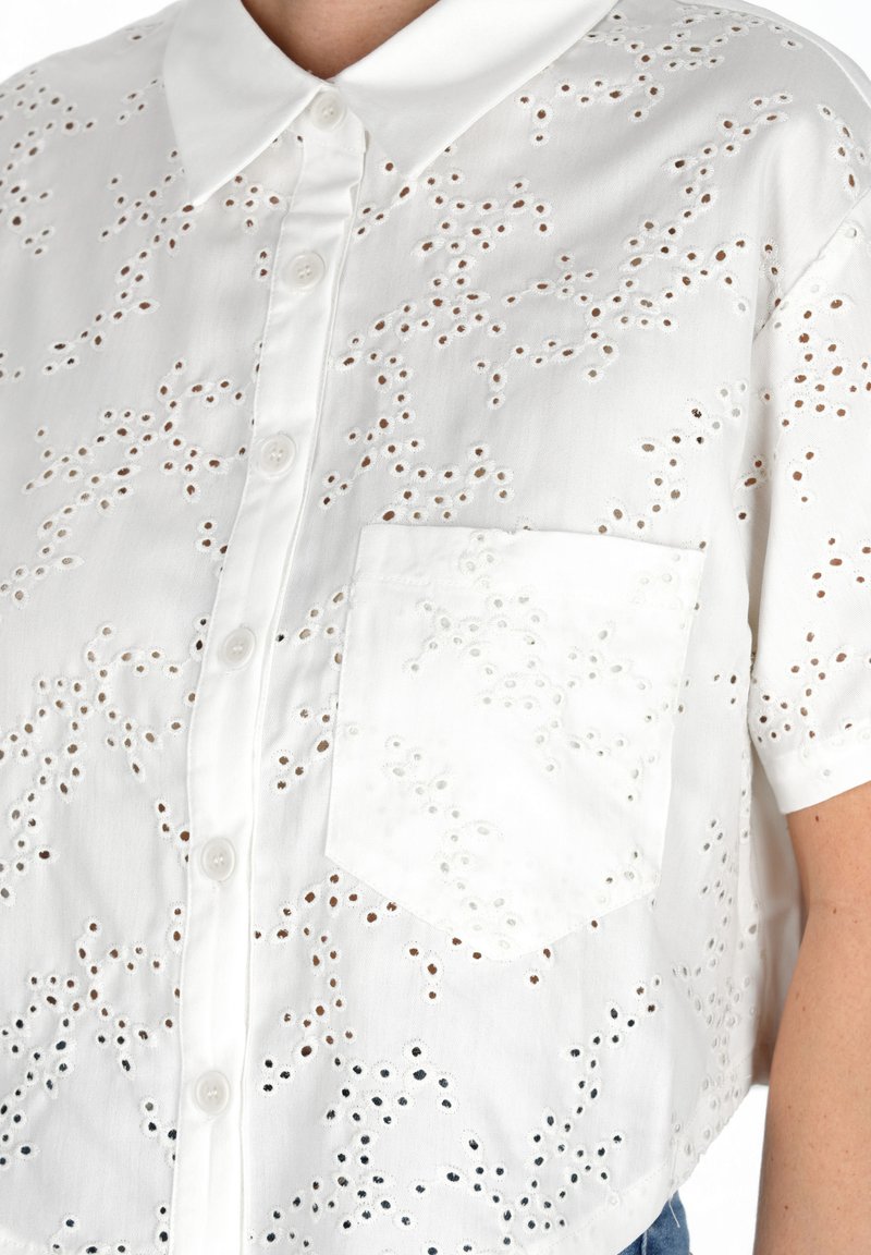 Camicia bianca in cotone con motivi ricamati a intaglio, maniche corte, colletto a punta e taschino sul lato sinistro.