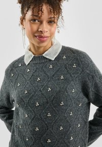 Donna con capelli ricci che indossa un maglione grigio scuro decorato con piccoli strass e una camicia bianca trasparente con colletto sotto.