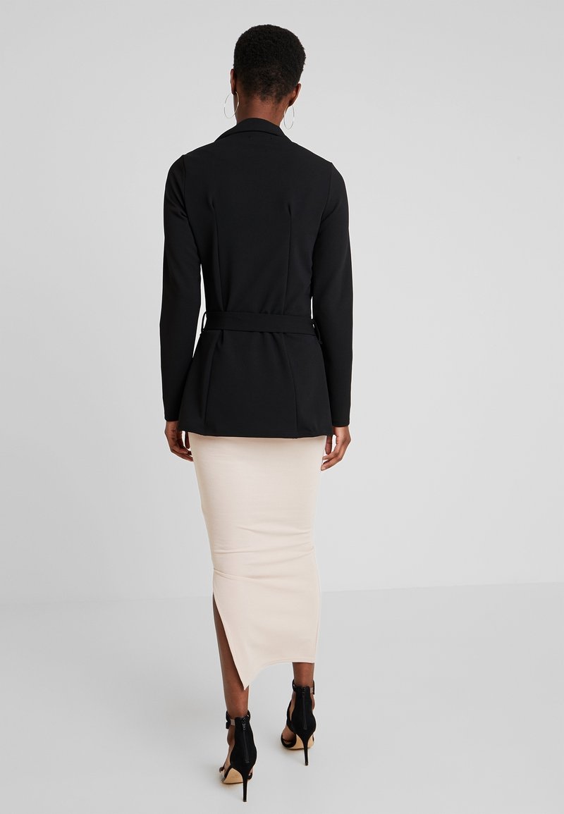 Czarna, dopasowana marynarka z długimi rękawami i paskiem w talii, noszona na jasnobeżową spódnicę ołówkową z bocznym rozcięciem oraz czarnymi szpilkami.