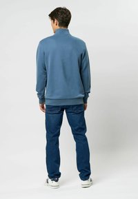 Kék pulóver magas gallérral, sötét kék farmernadrággal és fehér tornacipővel párosítva. Az anyag simának tűnik, bordázott mandzsettákkal és szegéllyel.