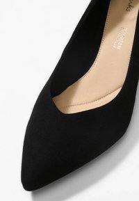 Chaussure plate noire à bout pointu avec texture en daim et semelle intérieure beige rembourrée.