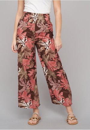 Kvinde iført bukser med vide ben med en brun base og pink, beige tropisk blomsterprint, kombineret med beige flade sandaler og en hvid top.