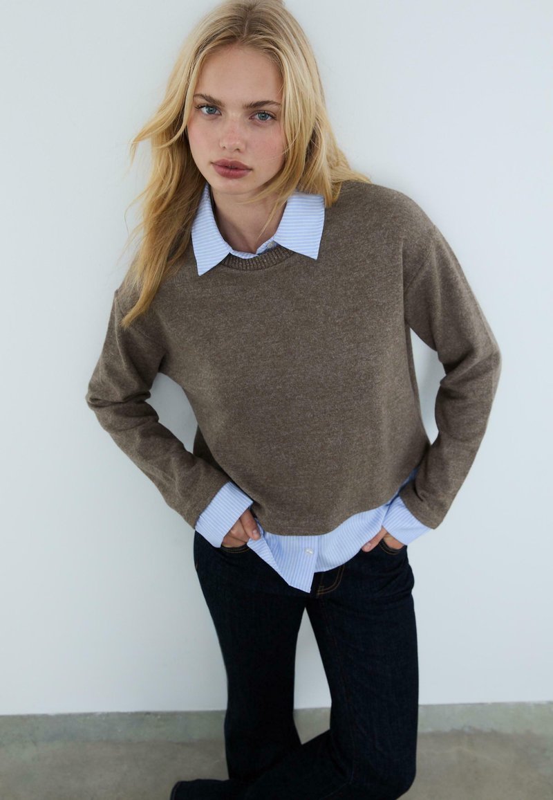 Brauner Strickpullover, der über einem hellblauen gestreiften Hemd mit Knöpfen getragen wird, mit rundem Ausschnitt und langen Ärmeln, kombiniert mit dunklen Jeans.