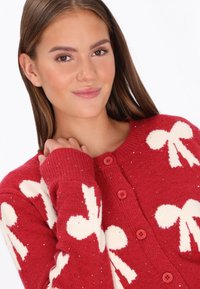 Fiatal nő piros kötött kardigánt visel, fehér masni mintákkal és piros gombokkal, lágyan mosolyogva, a gallérját simogatva.