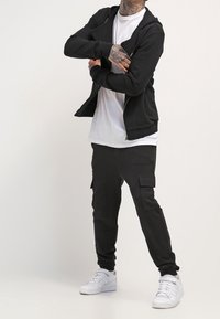 Sudadera negra con cremallera, camiseta blanca y pantalones cargo negros con bolsillos. Zapatillas blancas completan el conjunto, que presenta un ajuste relajado y un diseño sencillo.