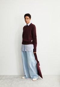 Bordeaux brei-sweater over een lichtblauw overhemd met kraag, gecombineerd met wijde lichtdenim broek. Houdt een bordeaux handtas vast.