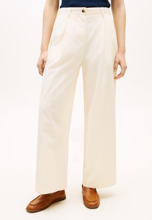 Frau trägt cremefarbene, weit geschnittene Hosen mit Falten, ein Marineblaues Oberteil und braune Ledermokassins, sie steht vor einem neutralen Hintergrund.