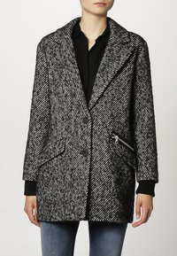 Abrigo de patrón de espiga en gris y negro con cuello con muescas, dos bolsillos frontales y un detalle de bolsillo con cremallera. La textura de la tela parece gruesa.