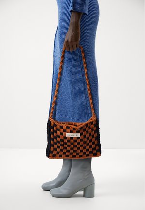 Personne en robe bleue côtelée et bottes grises tenant un sac tressé marron et noir étiqueté "Summery" sur un fond uni.