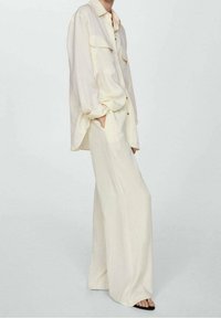 Losse crèmekleurige shirt met twee borstzakken, gecombineerd met wijde crèmekleurige broeken. Glad stoffen textuur en subtiele glans. Zwarte sandalen.