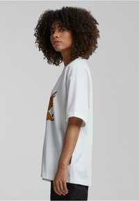 Hvit oversized T-skjorte med grafisk design som har en vinget dyrefigur, korte ermer og en avslappet passform. Stoffet ser mykt og teksturert ut.