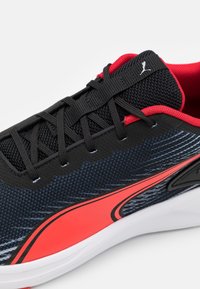 Černá a červená sportovní bota s texturovaným svrškem, síťovanými panely a bílou podrážkou; má logo na jazyku a designové akcenty na straně.