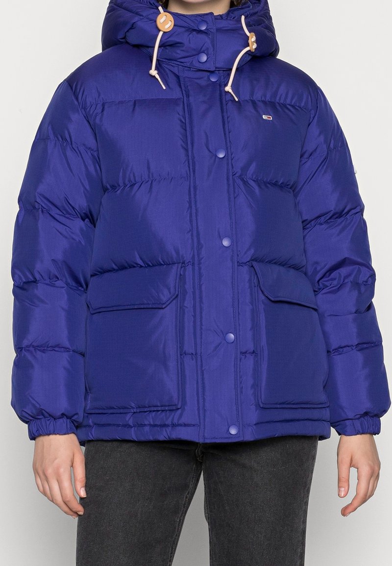 Veste matelassée bleue avec capuche, boutons-pression à l'avant, deux grandes poches à rabat et poignets élastiques, portée avec un jean foncé.