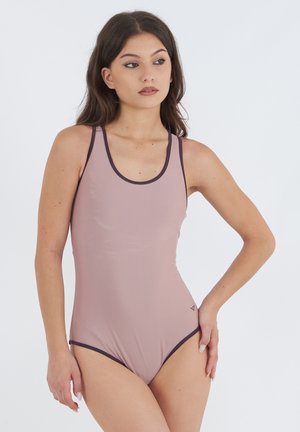 Jeune femme aux longs cheveux bruns portant un maillot de bain une pièce mauve avec des bordures foncées, debout avec une main sur la hanche devant un fond blanc.