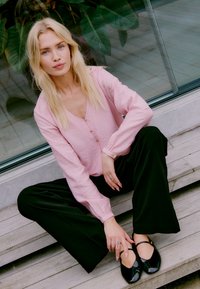 Blouse rose clair à manches longues avec un col en V et des détails de boutons, associée à un pantalon noir à jambes larges et des chaussures plates noires avec des brides de cheville.