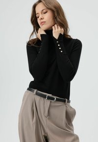 Zwarte coltrui met lange mouwen en knoopdetails, gecombineerd met licht beige geplooide broek en een zwarte riem.