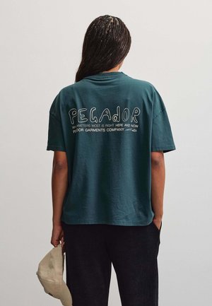 Person med fletninger bærer en mørkegrøn T-shirt med hvid tekst på ryggen og holder en beige kasket i venstre hånd.