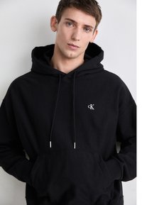 Čierna bavlnená mikina s kapucňou a predným vreckom, so šnúrkami na stiahnutie, ktorá má malé biele logo na hrudi. Hladký povrch a voľný strih.