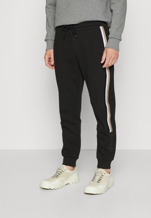 Schwarze Jogginghosen mit weißen, beigen und braunen Seitenstreifen, Kordelzug in der Taille, elastische Bündchen am Beinabschluss, getragen mit grauem Sweatshirt und beigen Sneakers.