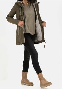 Veste isolante vert olive avec une fermeture éclair, une doublure grise et une capuche, associée à une chemise grise, un jean noir et des bottines couleur tan.