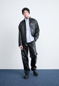 Svart fuskeskinnjakke og bukser kombinert med en lys blå skjorte med knapper. Outfiten har en teksturert strikket lue og kraftige svarte sko.