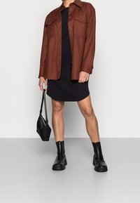 Veste marron avec deux poches poitrine, portée sur une robe noire ajustée. Des bottines noires et un petit sac à main noir complètent la tenue.
