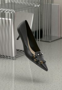 Černá kožená vysoká podpateková bota s špičatou špičkou, se zlatými a černými sponami a texturovaným povrchem. Umístěná v moderním prostředí.