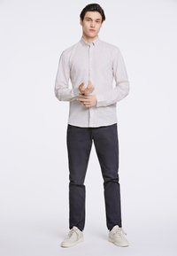 Cămașă bej deschis cu nasturi, realizată dintr-un material texturat, asortată cu pantaloni navy închis și adidași albi. Modelul stă într-o poziție neutră.