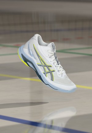 Pantof sport alb și gri Asics cu accente galbene suspendat deasupra unei podele de sală marcată cu linii colorate de teren.