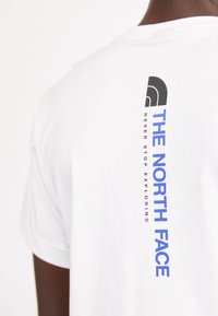Vit bomull t-shirt med "THE NORTH FACE" logotyp och texten "NEVER STOP EXPLORING" tryckt vertikalt i svart och blått på baksidan.