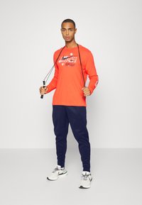 Longsleeve narančasta majica s bijelim grafičkim printom, u kombinaciji s tamnoplave udobne hlače. Model drži užad za preskakanje i nosi bijele sportske tenisice.
