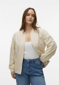 Beige bomber jacket with a zip front, ribbed cuffs, and relaxed fit, paired with blue high-waisted denim jeans and a white tank top.
