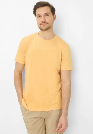 Mann trägt ein schlichtes gelbes kurzärmeliges T-Shirt und beige Hose, steht mit einer Hand in der Tasche vor weißem Hintergrund.