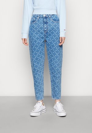 Persoon draagt een high-waist blauwe spijkerbroek met een diagonaal wit streeppatroon en witte sneakers, staand tegen een effen lichte achtergrond.