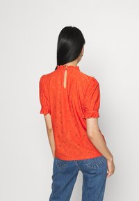Blusa rossa in tessuto leggero, con collo alto, maniche arricciate e retro con apertura a chiave. Sono visibili un motivo floreale e dettagli in rilievo.