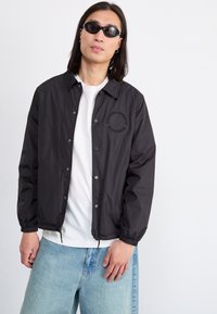 Black windbreaker jacket with snap buttons, featuring elastic cuffs and a logo print on the chest. Worn over a white shirt and blue jeans.