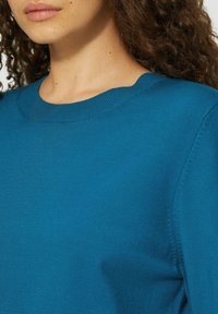 Turquoise trui met een ronde hals, zachte textuur, gladde stof en geribbelde details bij de kraag en de mouwranden.
