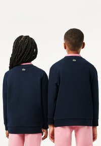 Des sweats bleu marine avec un col rose et un détail de logo vert, associés à un pantalon rose clair, vus de dos sur deux enfants.