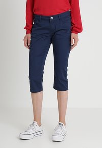 Pantalons cropped bleu marine en tissu doux, dotés d'un design à cinq poches, de passants de ceinture et d'une fermeture à bouton argenté. Portés avec des baskets blanches.