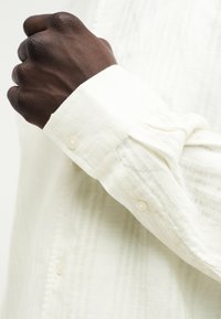 Ανοιχτόχρωμο πουκάμισο με υφή, μανσέτα με κουμπί και κάθετες ρίγες. Το ύφασμα φαίνεται απαλό και ελαφρώς διαφανές.