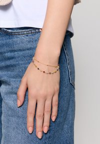 Bransoletka w kolorze złotym z kolorowymi koralikami w różnych rozmiarach, o delikatnym wzorze łańcuszka, noszona na dłoni na tle denimu.