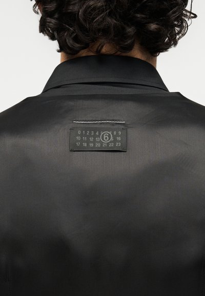Μαύρο πουκάμισο με απαλό υλικό, που διαθέτει μια χαρακτηριστική ετικέτα στο πίσω μέρος με αριθμούς και έναν κυκλικό λογότυπο. Κλασικός σχεδιασμός για το γιακά.