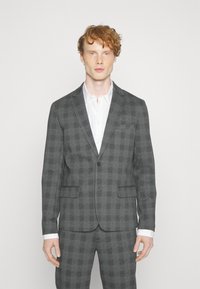 Grijs geruit pak met een enkele knoopsluiting, notch-kraag en twee voorzakken. Witte shirt eronder, met contrasterende manchetten zichtbaar.