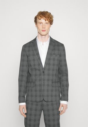 Grijs geruit pak met een enkele knoopsluiting, notch-kraag en twee voorzakken. Witte shirt eronder, met contrasterende manchetten zichtbaar.