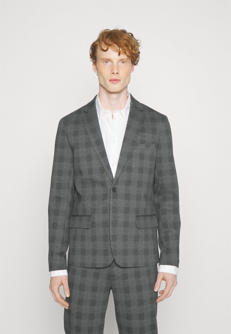 Grijs geruit pak met een enkele knoopsluiting, notch-kraag en twee voorzakken. Witte shirt eronder, met contrasterende manchetten zichtbaar.