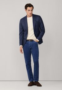 Blazer a cuadros azul marino superpuesto sobre un suéter crema, combinado con pantalones de pana azul y zapatos de ante marrón. Detalle sutil de pañuelo en el bolsillo.