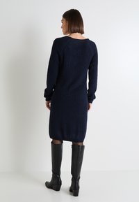 Vestido de punto azul marino con mangas largas y cuello redondo, que presenta un patrón texturizado. La modelo lleva botas de cuero negras hasta la rodilla.