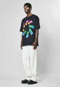 Černé oversized tričko s barevným, stylizovaným textem v různých písmech. Spárované s bílými kalhotami širokého střihu a černou obuví.