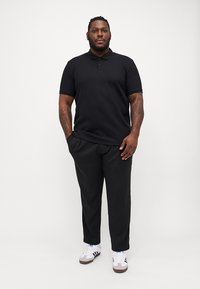 Sort polo-shirt med korte ærmer, båret med sorte bukser og hvide sneakers. Har en klassisk krave og en tætsiddende pasform.